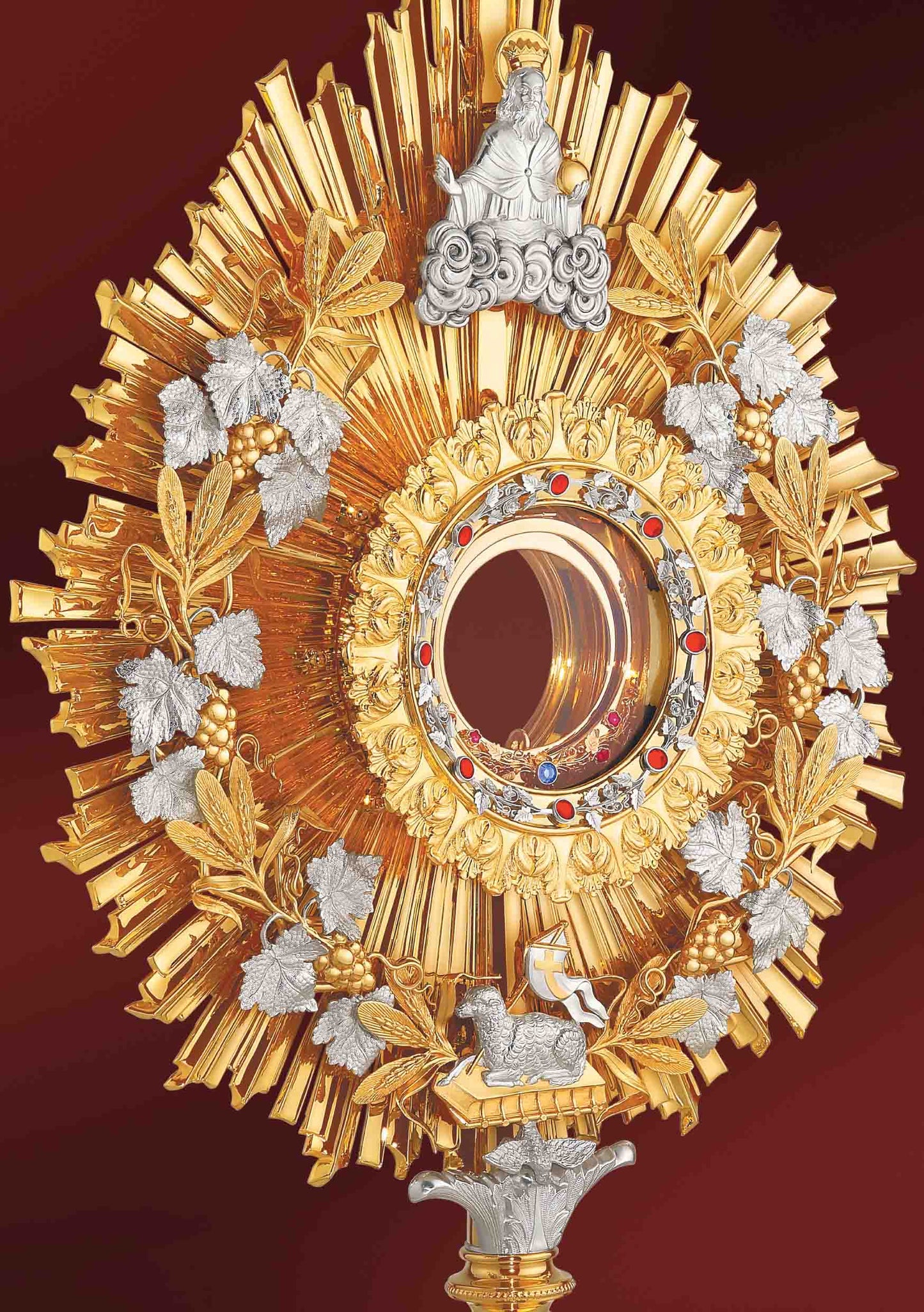 Eclectic brass monstrance, 24-karat gold plated, in the lightly pointed form topped by a cross. A wreath of grapevine leaves and crops adorns the sunburst glory. There are images of God the Father at the top and the Lamb of God at the bottom of the wreath. Equipped with a round luna graced with a floral motif. A decorative pattern of leaves set with red stones surrounds the luna case. Both the base and the nodus are richly embellished.