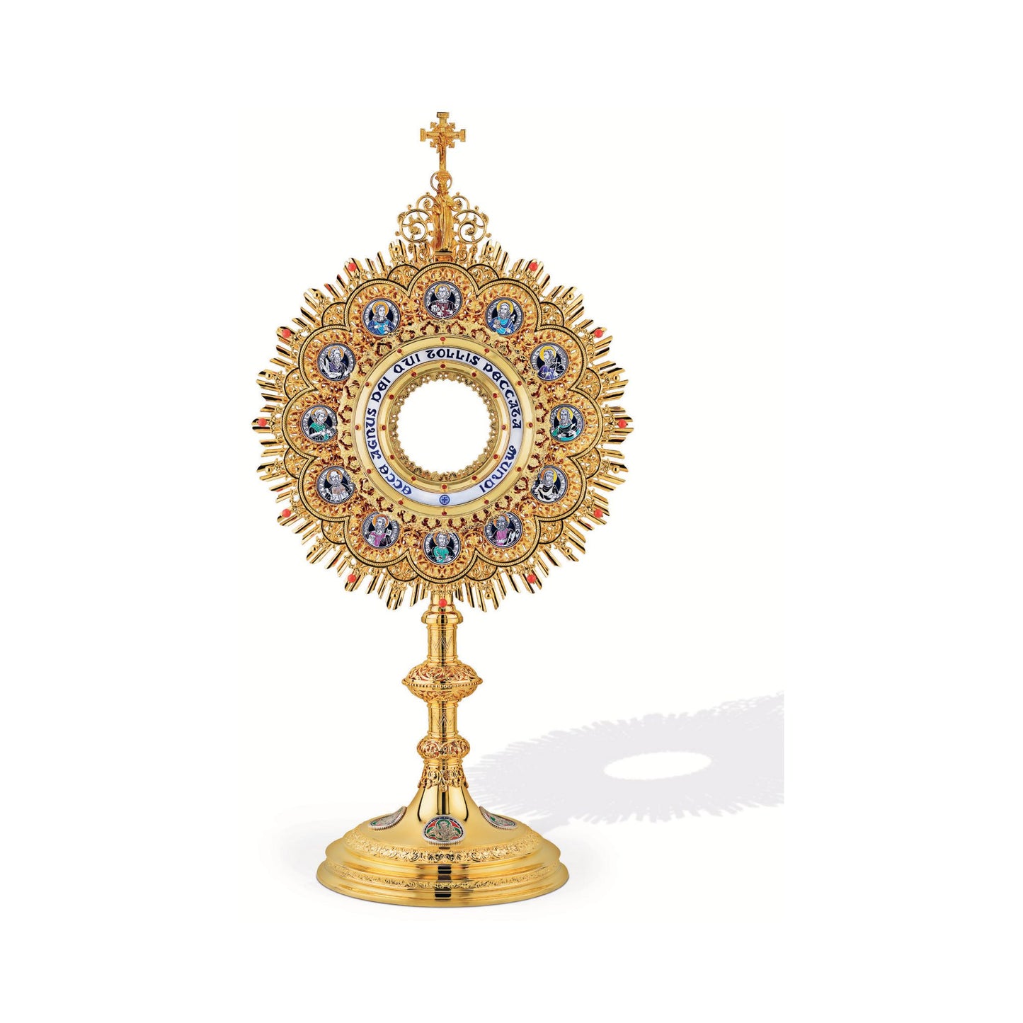 Brass monstrance, 24-karat gold-plated with red stones and enameled accents, inspired by German models from the late XIX and early XX centuries. The magnificent glory consists of a sunburst and a layer of ornate open filigrees with medallions depicting the Apostles. The gold-rimmed luna case has a blue-enameled Latin inscription: “Ecce Agnus dei Qui Tollis Peccata Mundi”. The stem and the node are decorated with fine filigrees. Medallions on the round base feature symbols of the Evangelists. All medallions
