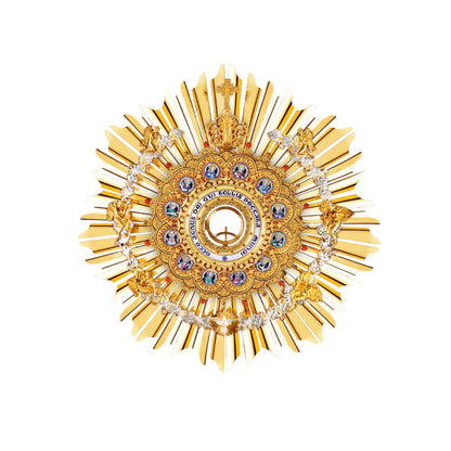 Brass hanging monstrance, 24-karat gold-plated with red stones and enameled accents, inspired by German models from the late XIX and early XX centuries. The magnificent glory consists of a sunburst and a layer of ornate open filigrees with medallions depicting the Apostles. The gold-rimmed luna case has a blue-enameled Latin inscription: “Ecce Agnus dei Qui Tollis Peccata Mundi”. The stem and the node are decorated with fine filigrees. Medallions on the round base feature symbols of the Evangelists. All med