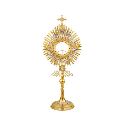 A large monstrance (height: 43-1/4") for a big host (diameter: 6"). Made of brass, 24-karat gold-plated. The glory consists of two sunbursts and two elaborate silver-plated wreaths. The bigger wreath has figures of God the Father and Jesus attached to the top, and six angels placed around the luna case. The smaller wreath is embellished with blue-enameled accents. A figure of the Holy Spirit (the Dove) against its own small sunburst is set at the bottom of the glory. 