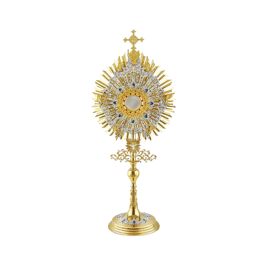 A large monstrance (height: 43-3/4") made of brass, 24-karat gold-plated with silver-plated and blue-enameled accents. The sunburst-shaped glory consists of three layers of rays and two silver-plated wreaths made of ornate floral scrollwork. The bigger wreath is adorned with figures of God the Father and Jesus at the top, and six angels around the luna case. The opening for the luna is gold-rimmed, filigreed and encrusted with synthetic red gemstones. A figure of the Holy Spirit (the Dove) is placed at the 