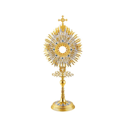 A large monstrance (height: 43-3/4") made of brass, 24-karat gold-plated with silver-plated and blue-enameled accents. The sunburst-shaped glory consists of three layers of rays and two silver-plated wreaths made of ornate floral scrollwork. The bigger wreath is adorned with figures of God the Father and Jesus at the top, and six angels around the luna case. The opening for the luna is gold-rimmed, filigreed and encrusted with synthetic red gemstones. 