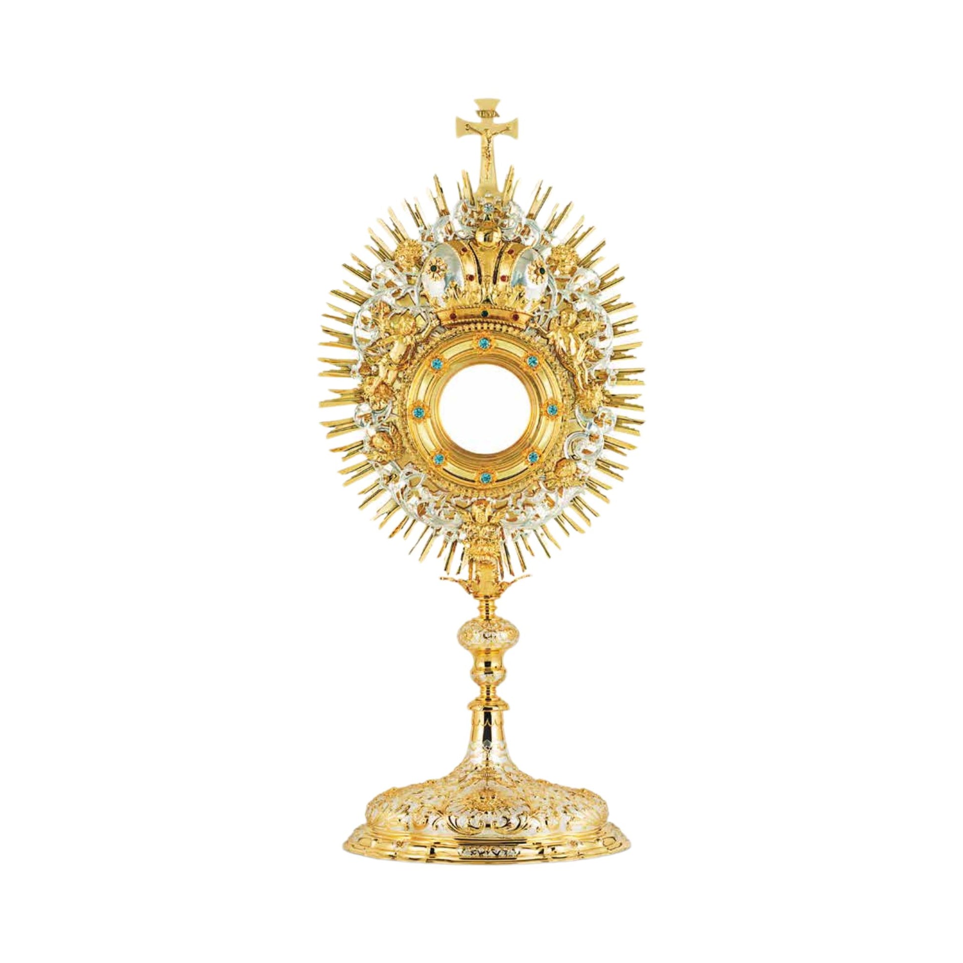 Baroque monstrance made of brass, 24-karat gold-plated with silver-plated accents. The luna case is encrusted with synthetic blue stones and surrounded by a decorative wreath with a crown and images of angels. The glory is sunburst-shaped and topped with a cross. The node and the base are adorned with rich ornamental patterns in two tones. 