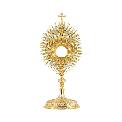 Baroque monstrance made of brass, 24-karat gold-plated with silver-plated accents. The luna case is encrusted with synthetic blue stones and surrounded by a decorative wreath with a crown and images of angels. The glory is sunburst-shaped and topped with a cross. The node and the base are adorned with rich ornamental patterns in two tones. 