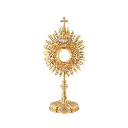Baroque monstrance made of brass, 24-karat gold plated with silver plated accents. The glory consists of a sunburst topped with a cross and a decorative layer of floral ornaments around the luna case. God the Father and the Holy Spirit are placed at the top of the glory, while the Lamb of God is placed at the bottom. On either side of the luna case there is an angel figure. Both the node and the oval base richly adorned with floral ornaments and images of angels. The monstrance is coated with a protective l