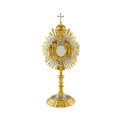 Baroque brass monstrance, gold-plated with silver-plated detailing. The central part consists of a sunburst and a silver-plated wreath of wheat and grapevine. At the top of the opening for the luna there is an image of God the Father and the Dove (the Holy Spirit). The Lamb of God is attached to the bottom of the glory. Several angel figures surround the luna case. The monstrance is topped with a canopy and a crucifix with a silver-plated figure of Jesus. The base is embellished with silver-plated and oxidi