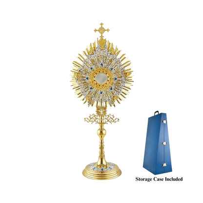 A large monstrance (height: 43-3/4") made of brass, 24-karat gold-plated with silver-plated and blue-enameled accents. The sunburst-shaped glory consists of three layers of rays and two silver-plated wreaths made of ornate floral scrollwork. The bigger wreath is adorned with figures of God the Father and Jesus at the top, and six angels around the luna case. The opening for the luna is gold-rimmed, filigreed and encrusted with synthetic red gemstones. A figure of the Holy Spirit (the Dove) is placed at the 