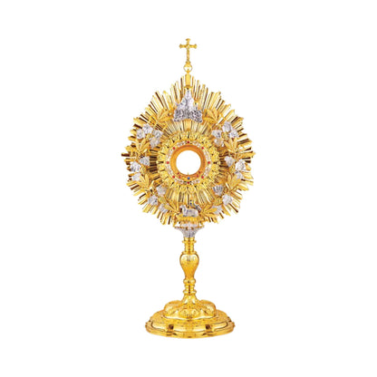 Eclectic brass monstrance, 24-karat gold plated, in the lightly pointed form topped by a cross. A wreath of grapevine leaves and crops adorns the sunburst glory. There are images of God the Father at the top and the Lamb of God at the bottom of the wreath. Equipped with a round luna graced with a floral motif. A decorative pattern of leaves set with red stones surrounds the luna case. Both the base and the nodus are richly embellished.