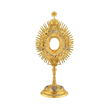 Brass monstrance, 24-karat gold-plated with silver-plated accents. The glory is embellished with an ornate wreath featuring grapevine and wheat motifs, and figures of God the Father, two angels and the Ark of The Covenant. The monstrance is topped with a dome and a cross encrusted with a pink stone. The oval base is decorated with silver-plated medallions of the Evangelists.